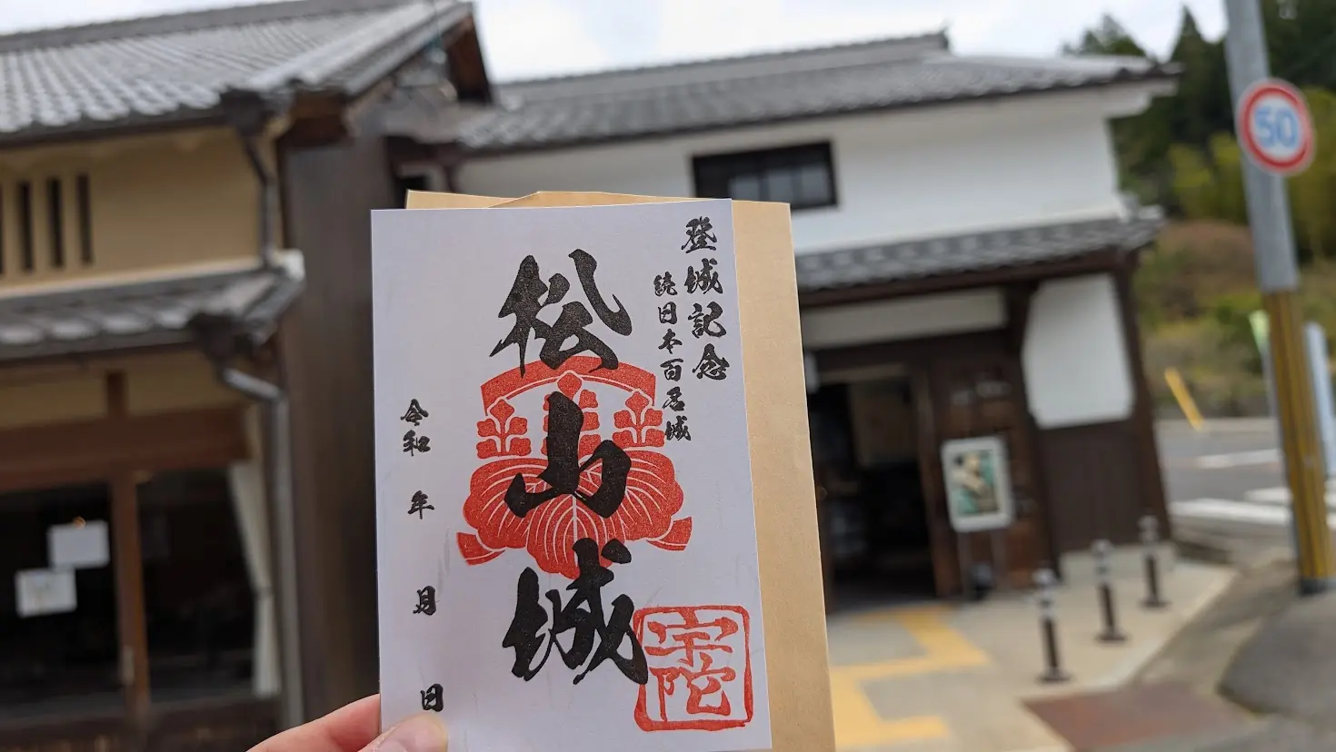宇陀松山城跡の御城印。「登城記念 続日本百名城 松山城」と記され、赤い家紋と宇陀の朱印が押されている。背景には宇陀松山の歴史的な町並みが写っている。