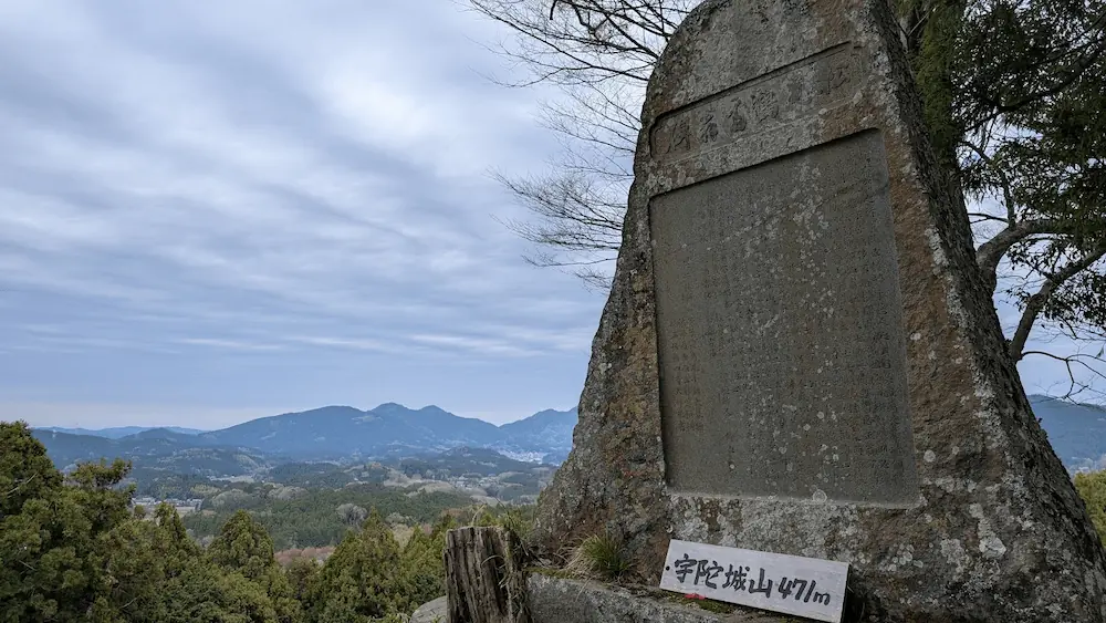 宇陀松山城跡の頂上に立つ大きな石碑。足元には「宇陀城山 471m」と書かれた札があり、背景には宇陀の山々と広い空が広がっている。