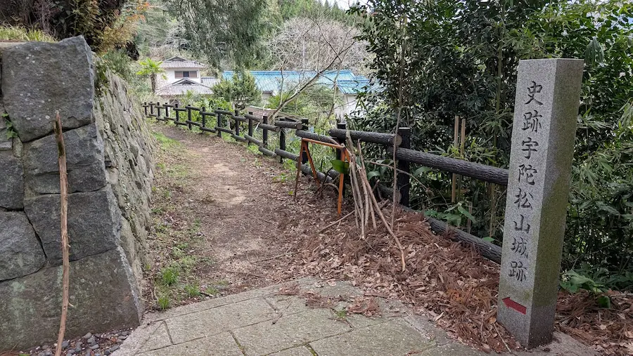 奈良県宇陀市・春日神社の脇にある「史跡 宇陀松山城跡」の登城路入り口。石造りの標柱に赤い矢印があり、左側には石垣、奥には木製柵が続く未舗装の山道が伸びている。