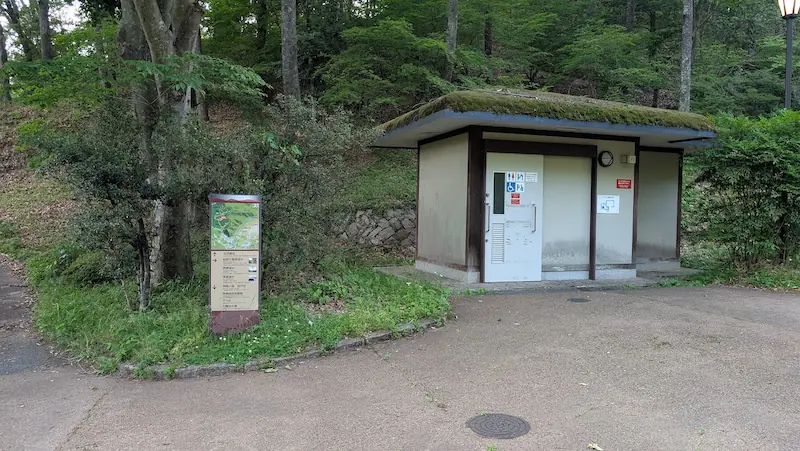 明日香村のハイキングコース中腹に設置された、屋根に苔が生した公衆トイレと、その横に立つ観光案内板。