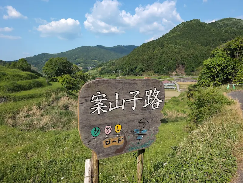 明日香村稲渕の「案山子路(かかしロード)」を示す木製の看板。背景には初夏の棚田と山々が広がっている