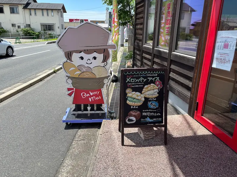 ベーカリーメイの入り口に設置されたパンを持つ子供のキャラクターパネルと、メロンパンアイスを紹介する手書きの黒板メニュー。