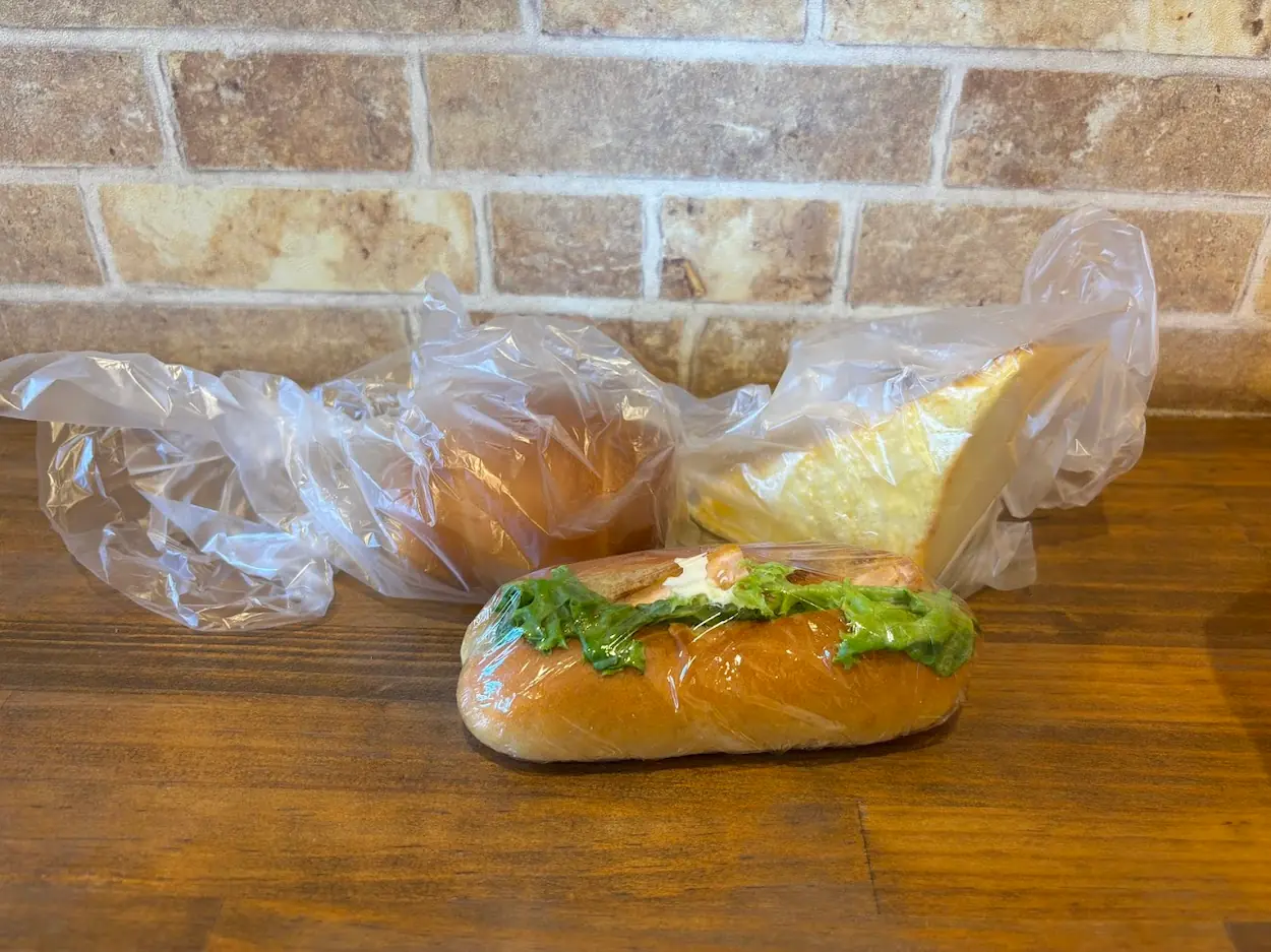 ベーカリーメイで購入した、塩パン、厚切りフレンチトースト、えびかつこっぺの3種類のパン。木製のテーブルの上に置かれている。