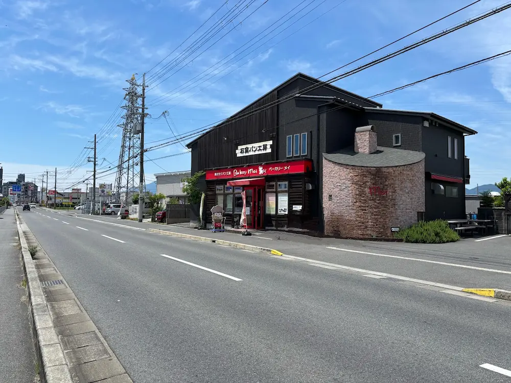 大和高田市の県道沿いに建つ黒い外壁の「ベーカリーメイ」外観と、店前を通る道路の全景。