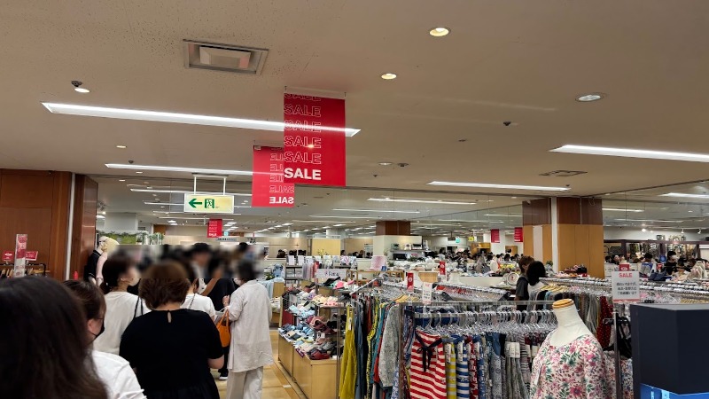 セール会場で多くの買い物客で賑わう店内の様子。頭上には赤い「SALE」のバナーが掲げられ、多くの人が列を作っている