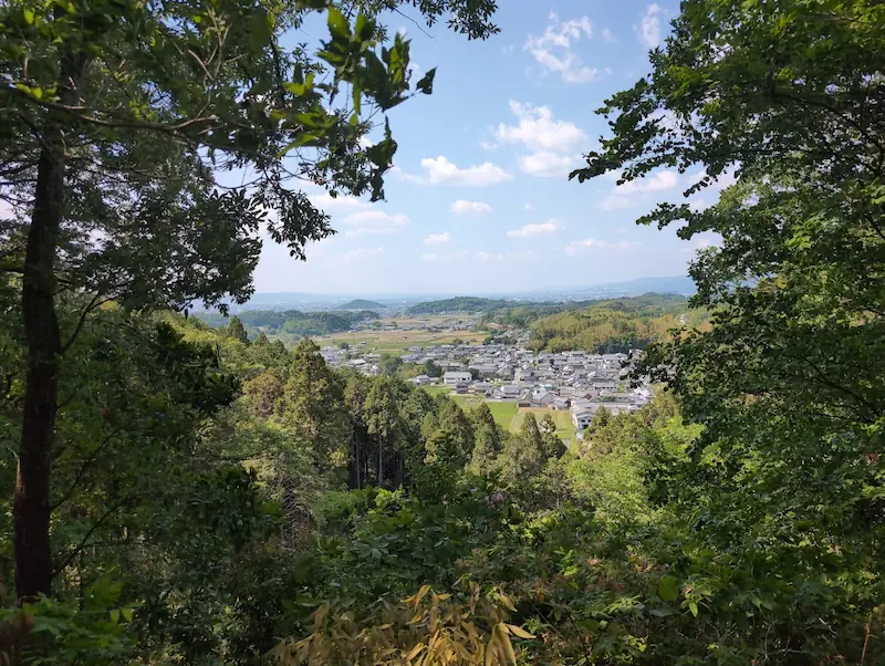 東展望台から木々の間越しに見下ろす明日香村の集落と田園風景。晴れた空の下、飛鳥の里が広がっている。