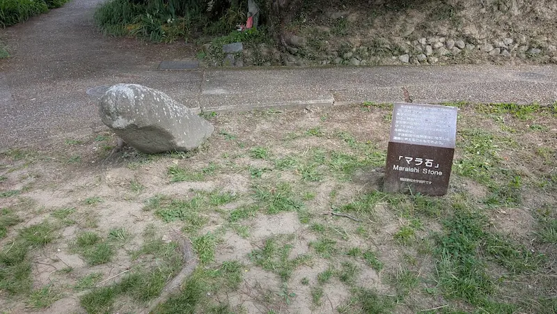 明日香村にある飛鳥時代の石造物「マラ石」。道路脇の地面に横たわる石と、その横に立つ解説看板。