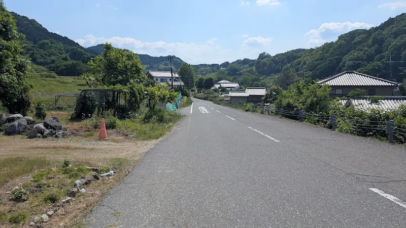 都塚古墳の近くから稲渕棚田方面へ続く、広くてなだらかな下り坂の舗装路。周囲には民家やのどかな田園風景が広がる。