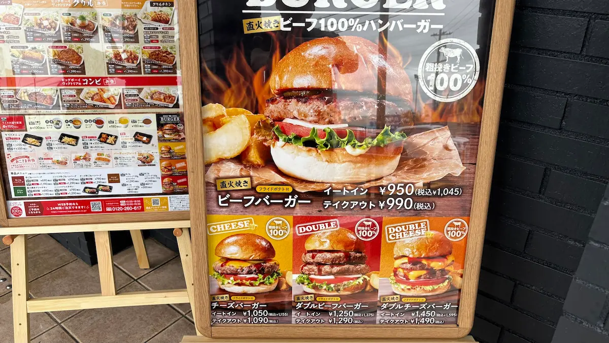 ビッグボーイの店頭にある、直火焼きビーフ100%ハンバーガーとテイクアウト弁当のメニュー看板。ビーフバーガー、チーズバーガー、ダブルチーズバーガーなどのラインナップと価格が記載されている