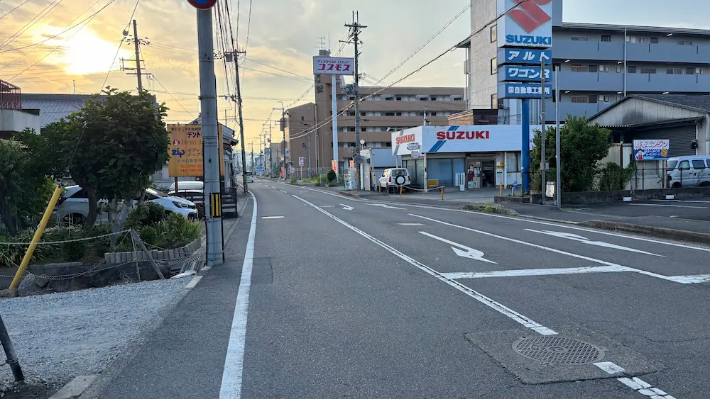 「BRACELET LAB.」の店舗付近から運転免許センター方面へ向かう道路の様子。夕暮れ時の光の中、右側にスズキの看板やディスカウントドラッグコスモス、左側に電柱や植栽が写っている。