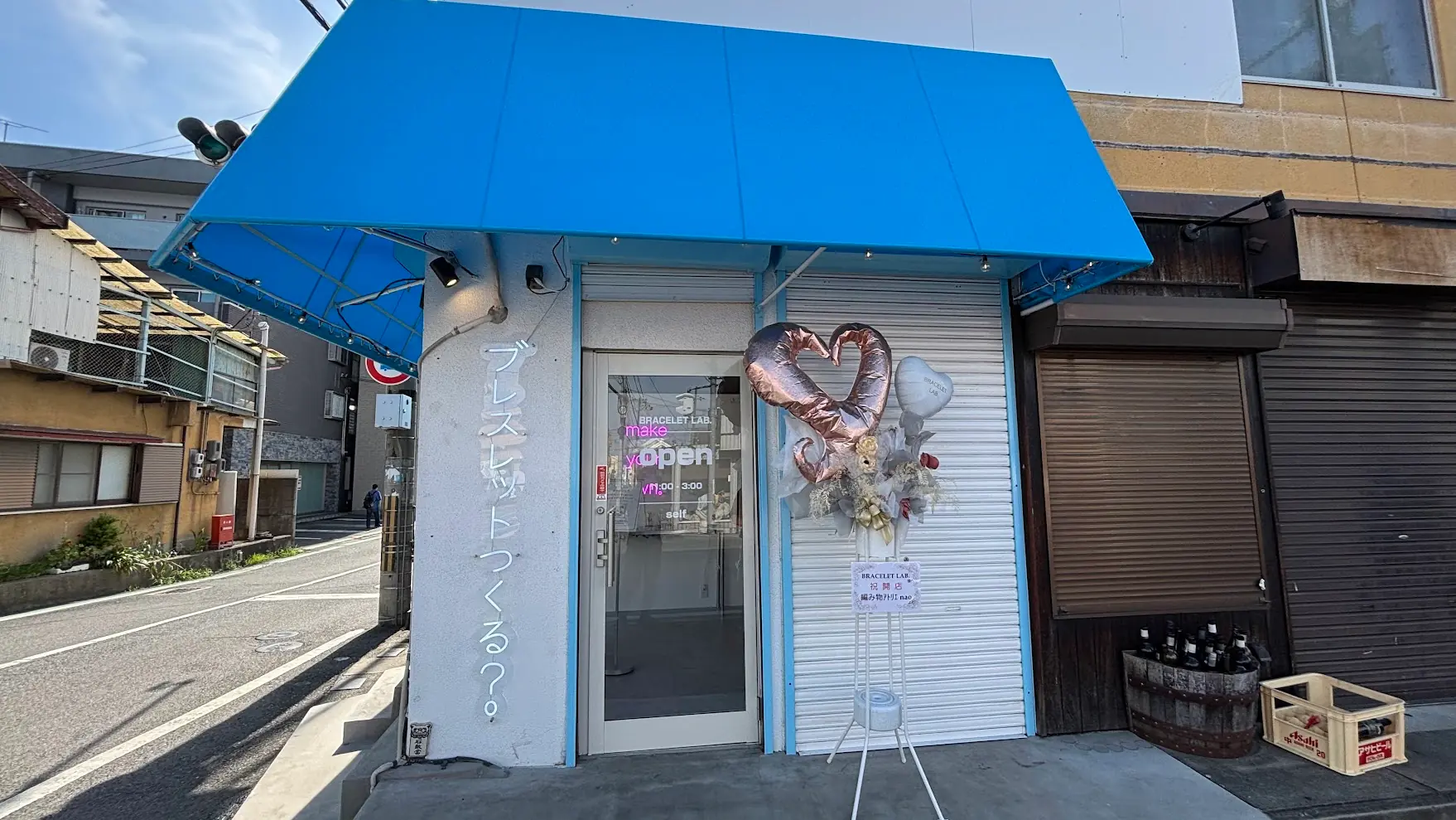 青い大きなオーニングテントが特徴的な「BRACELET LAB.（ブレスレットラボ）」の店舗外観。ガラス扉の横に開店祝いのハート型のバルーンスタンドが飾られている。