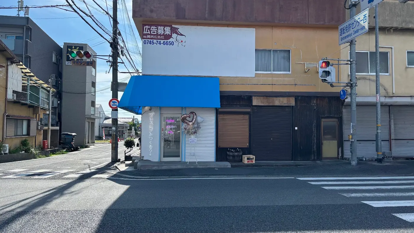 青いオーニングテントが特徴的な「BRACELET LAB.」の店舗外観と、交差点や周辺の道路、電柱、看板が写る周囲の様子。