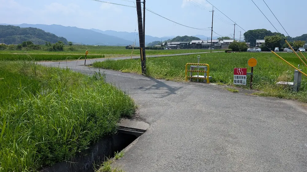 田園地帯を走る極めて狭いアスファルトの農道の脇に側溝があり、その奥に「花園見学者駐車場」の看板と駐車された車が見える様子。