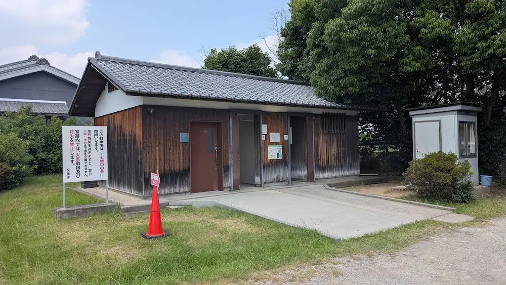 木目調の外壁に瓦屋根の落ち着いたデザインの藤原宮跡駐車場トイレの外観。手前に「この駐車場は閉門します」と書かれた立て看板と赤いコーンが置かれている。