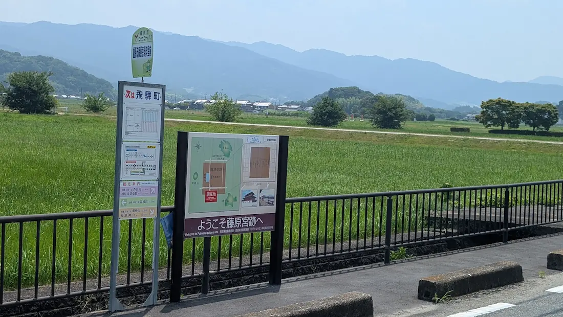 田園風景と山々を背景に、橿原市コミュニティバスの「橿原市藤原京資料室前」バス停と「ようこそ藤原宮跡へ」と書かれた周辺案内マップが設置されている様子。