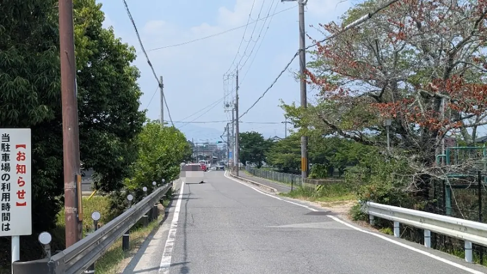藤原宮跡の前の道路の様子。片側1車線のアスファルト道路が奥へと伸びており、前方を走る車と遠くに信号機、左右に木々やガードレールが写っている。左手前には「お知らせ」と書かれた看板が掲げられている。