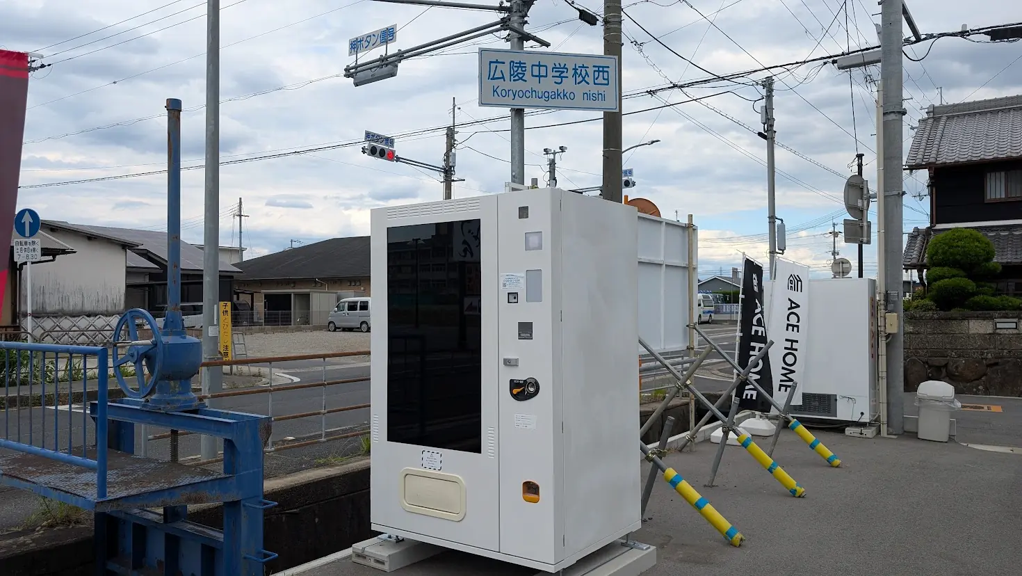 広陵中学校西交差点の近くに設置された、白い筐体の謎の自動販売機。周囲には工事用のフェンスや資材が見られ、近日オープン予定の雰囲気が漂っている