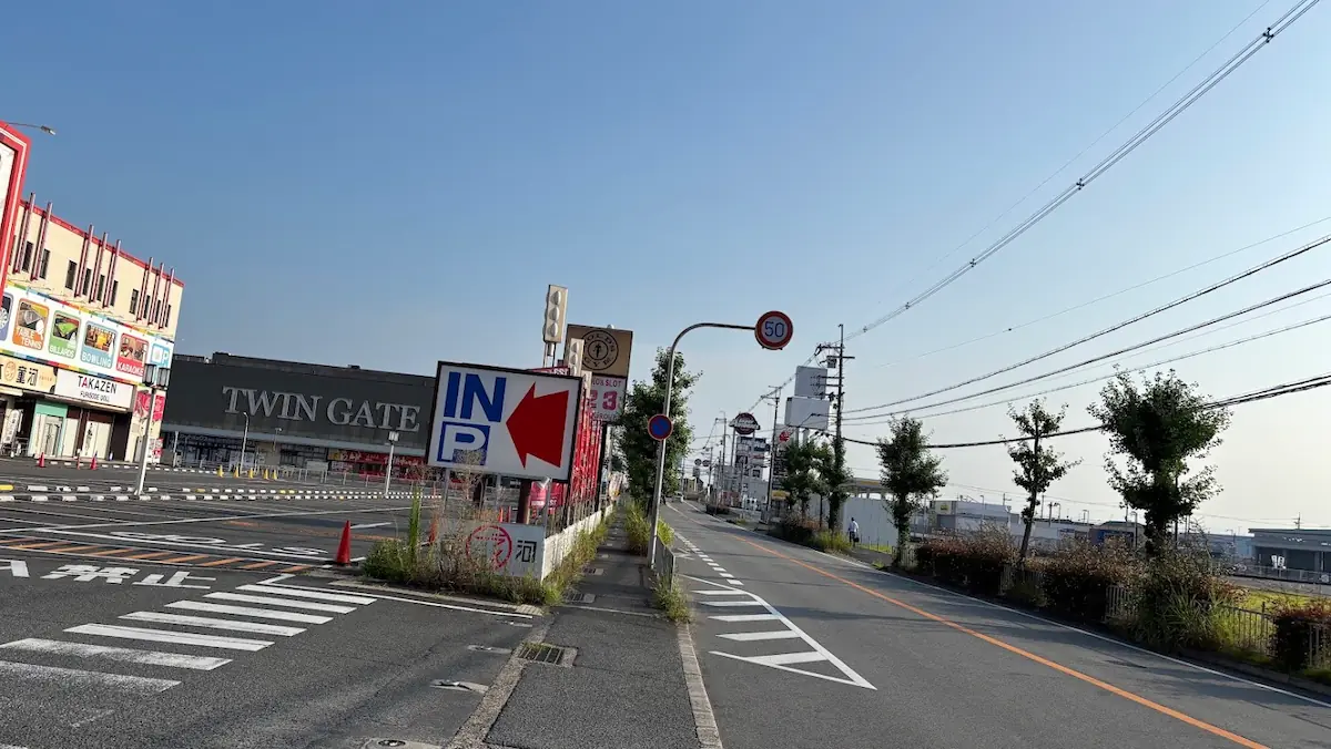 駐車場の入り口付近から奈良方面へ向かう道路の様子。左側にTWIN GATEの建物と駐車場、右側に街路樹が並ぶ片側1車線の道路が続いている。