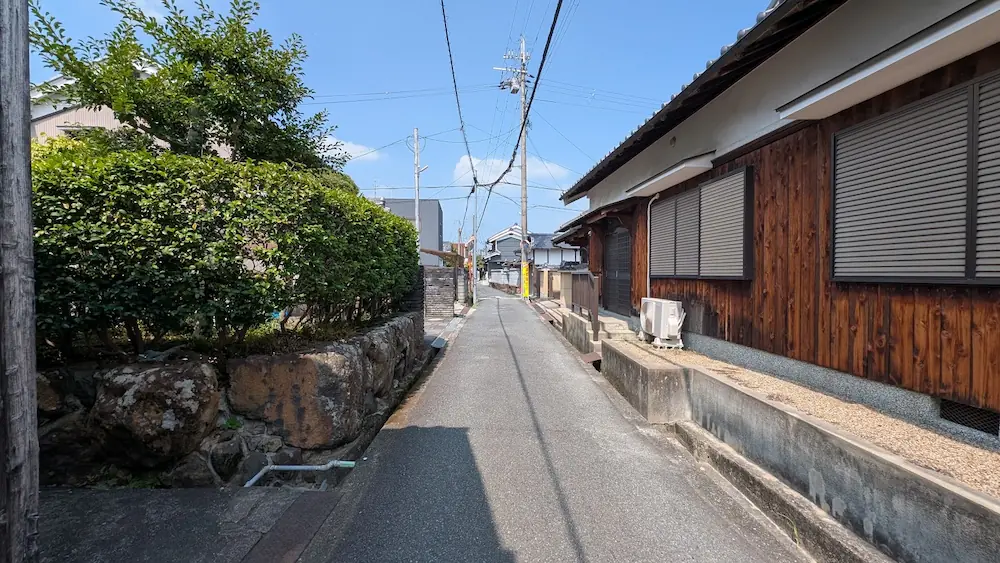 おふさ観音周辺の、車一台がやっと通れるような住宅街の狭いアスファルト道路の様子。左側に石垣と生け垣、右側に木造家屋が並んでいる。