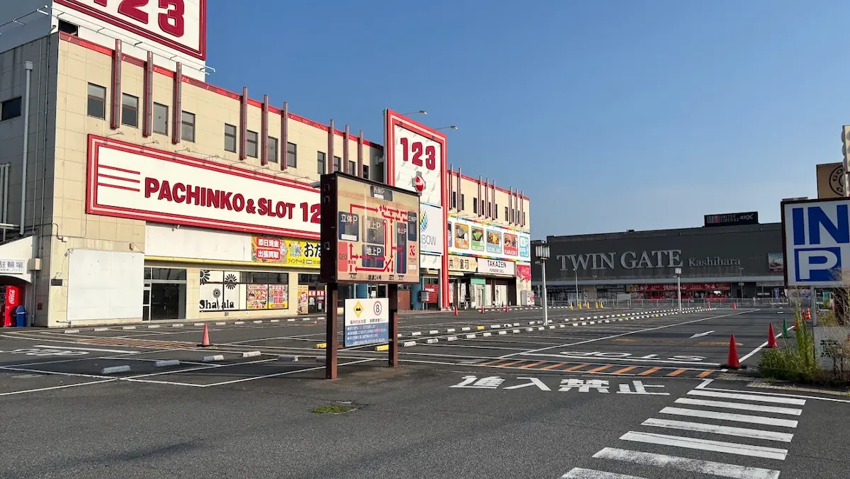 パチンコ店「123」や「TWIN GATE」の大きな建物に囲まれた、広々としたアスファルト舗装の駐車場と案内看板の様子。