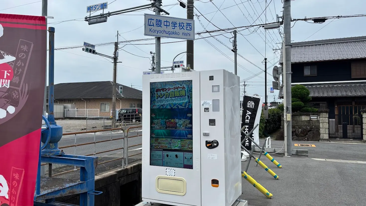 日中の「広陵中学校西」交差点。青空の下、白い「ポケモンカードゲーム トレカ自販機」が設置されており、周辺の道路や住宅、電柱が写っている