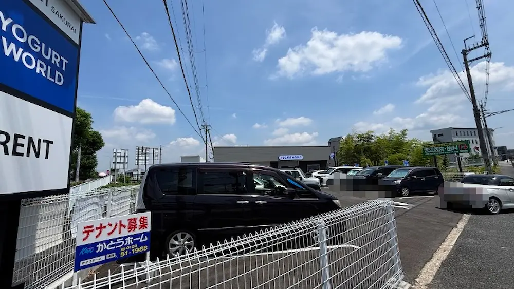本日グランドオープンを迎えた「桜井ヨーグルトワールド」の駐車場で、午前11時頃に多くの車が駐車され賑わっている様子。手前には「テナント募集」の看板と白いフェンスが写っている。