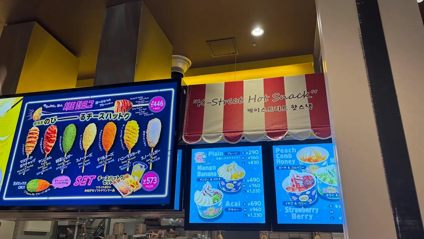 太陽のトマト麺橿原アルル店の店頭にある、チーズハットグとフローズンヨーグルトアイスのメニュー看板。7種類のフレーバーが選べるチーズハットグと、マンゴーやアサイーなどのトッピングが豊富なヨーグルトアイスが掲載されている