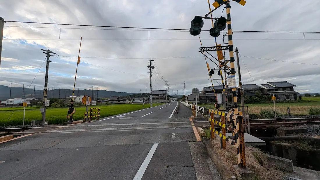 曇天の下、「Cafe ゆるり」付近の踏切から明日香方面へ向かって伸びる車道や周囲の田園風景を見渡した様子。