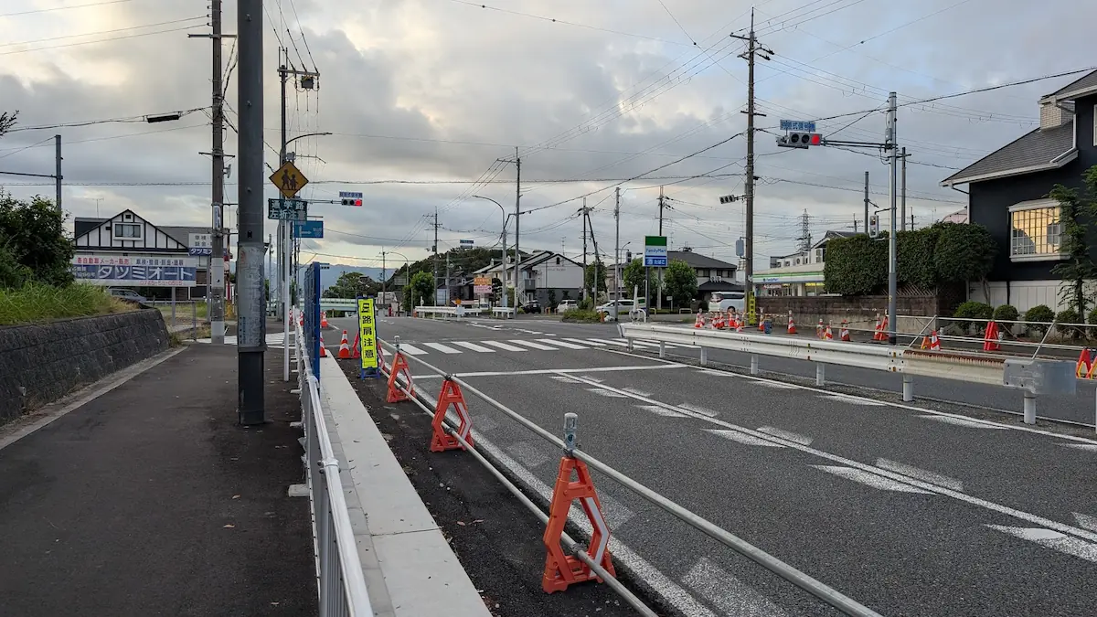 夕暮れの空の下、中和幹線の工事現場から高田方面へ向かう道路の様子。道路脇にはオレンジ色のカラーコーンや仮設フェンスが並び、前方に信号機や周辺の建物が見える。