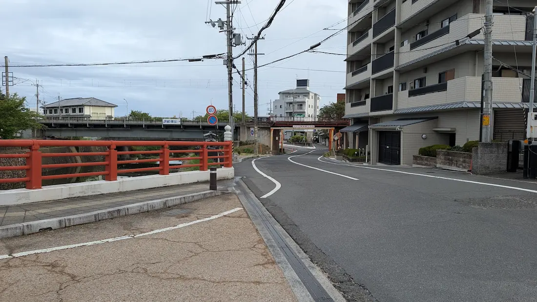 朱色の高欄が特徴的な橋を渡り、今井町方面へと続く道路と、その奥に見えるJRの鉄道高架や周辺の建物の様子。
