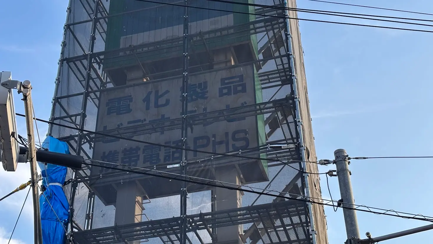 青空の下、足場と黒い養生メッシュに囲まれた橿原市の旧ダイソー看板の様子。「電化製品」などの文字がうっすらと見えている。