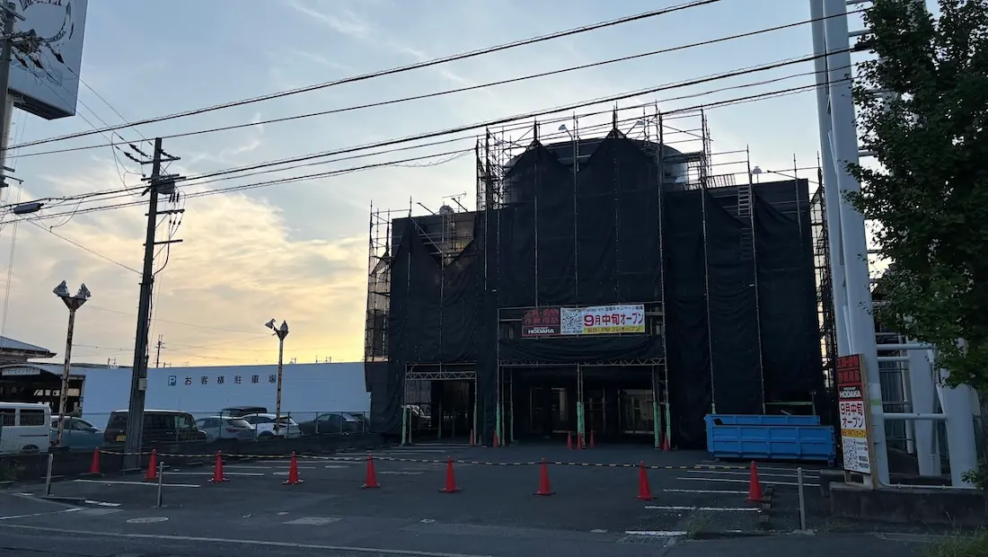 夕暮れの青空の下、全面に黒い足場メッシュが組まれ、9月中旬オープンを知らせる横断幕が掲げられた橿原市の「PRO SHOP HODAKA」の店舗外観。