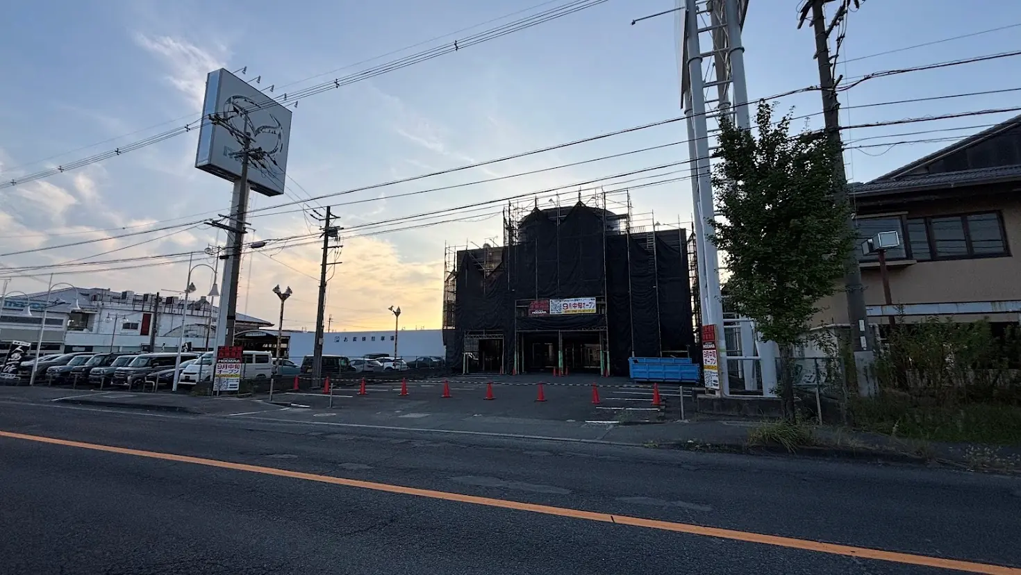 夕暮れの青空の下、左側にマツダの看板が立ち、正面に工事中の「HODAKA(ホダカ)」店舗と広々としたお客様駐車場が広がる道路沿いの景観。