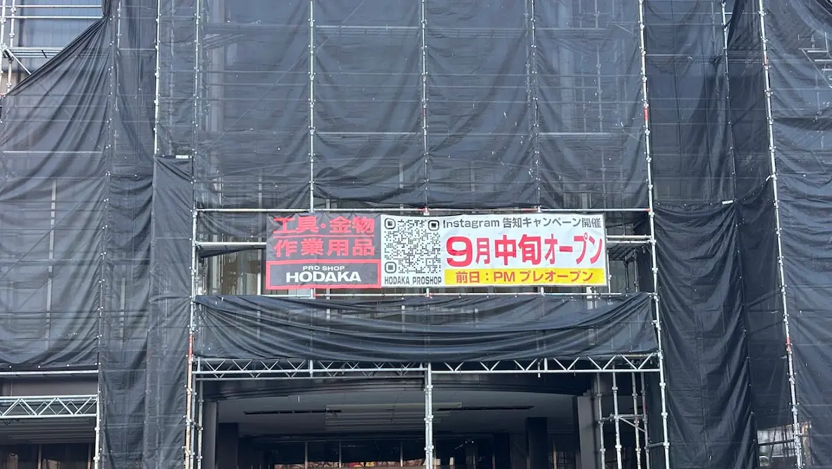 黒い足場メッシュに覆われた工事中の店舗外観に掲げられた、「9月中旬オープン」「前日:PMプレオープン」および工具・金物・作業用品の「PRO SHOP HODAKA」とQRコードが記載された横断幕。