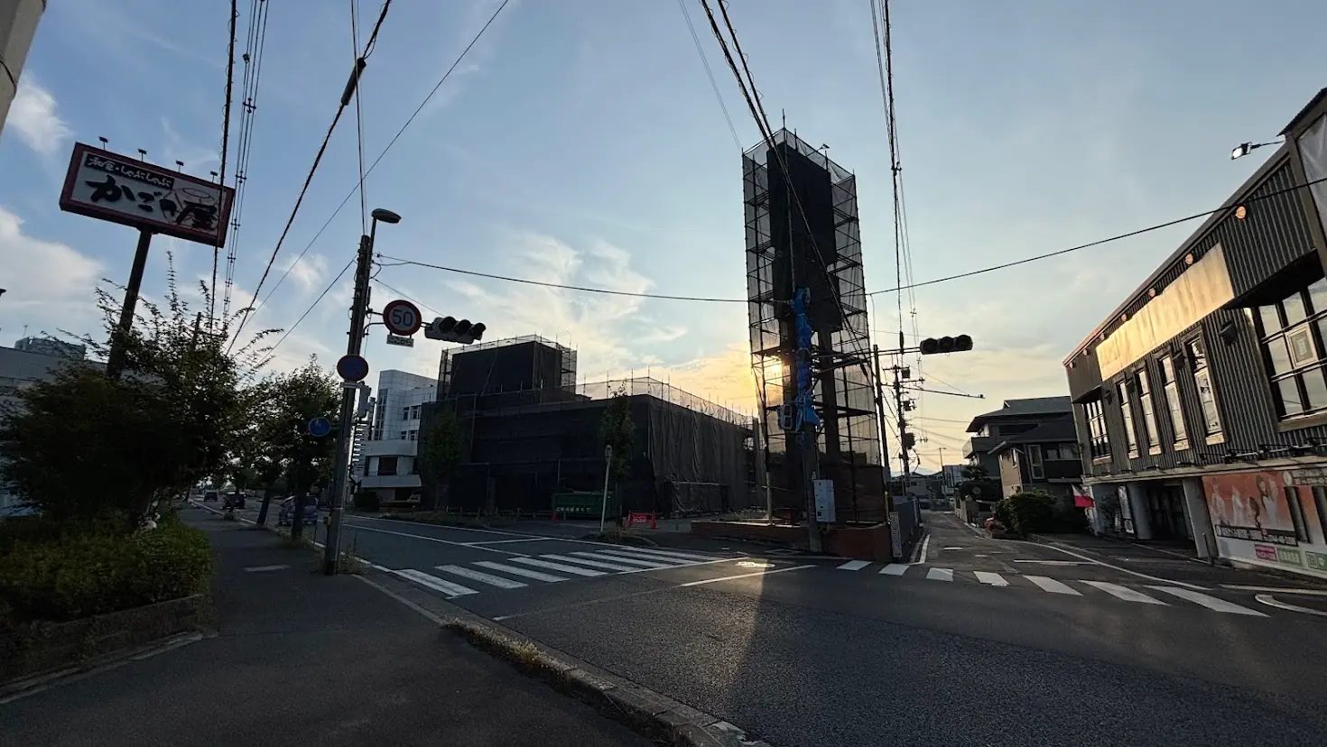 夕暮れの青空の下、左側に「かごの屋」の赤い看板、正面に足場に覆われた工事中の建物や道路が写る橿原市内の交差点付近の様子。