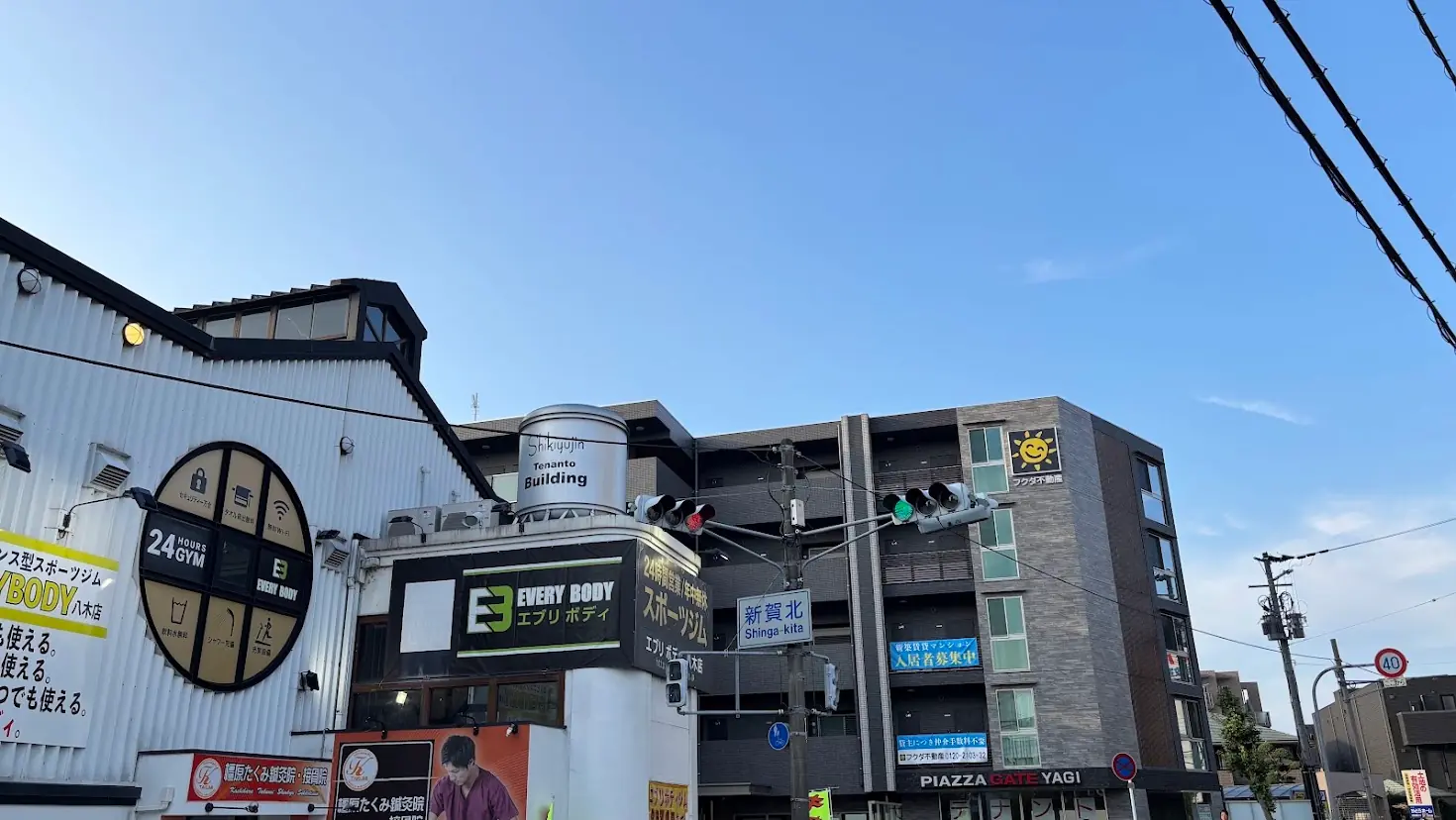 青空の下、奈良県橿原市にある「新賀北」交差点付近の様子。エブリボディ八木店などのテナントビルや交差点名を示す青い標識が写っている。