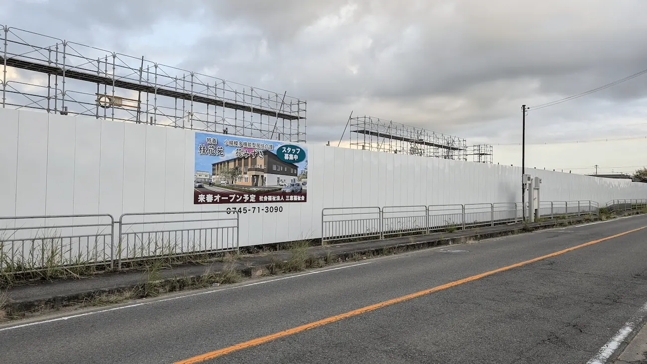 夕暮れの空の下、県道5号線沿いに続く白い仮囲いと、「特養 桂花苑」「花みずき」の完成予想図看板、および上部に組まれた足場全体を捉えた建設現場の様子。
