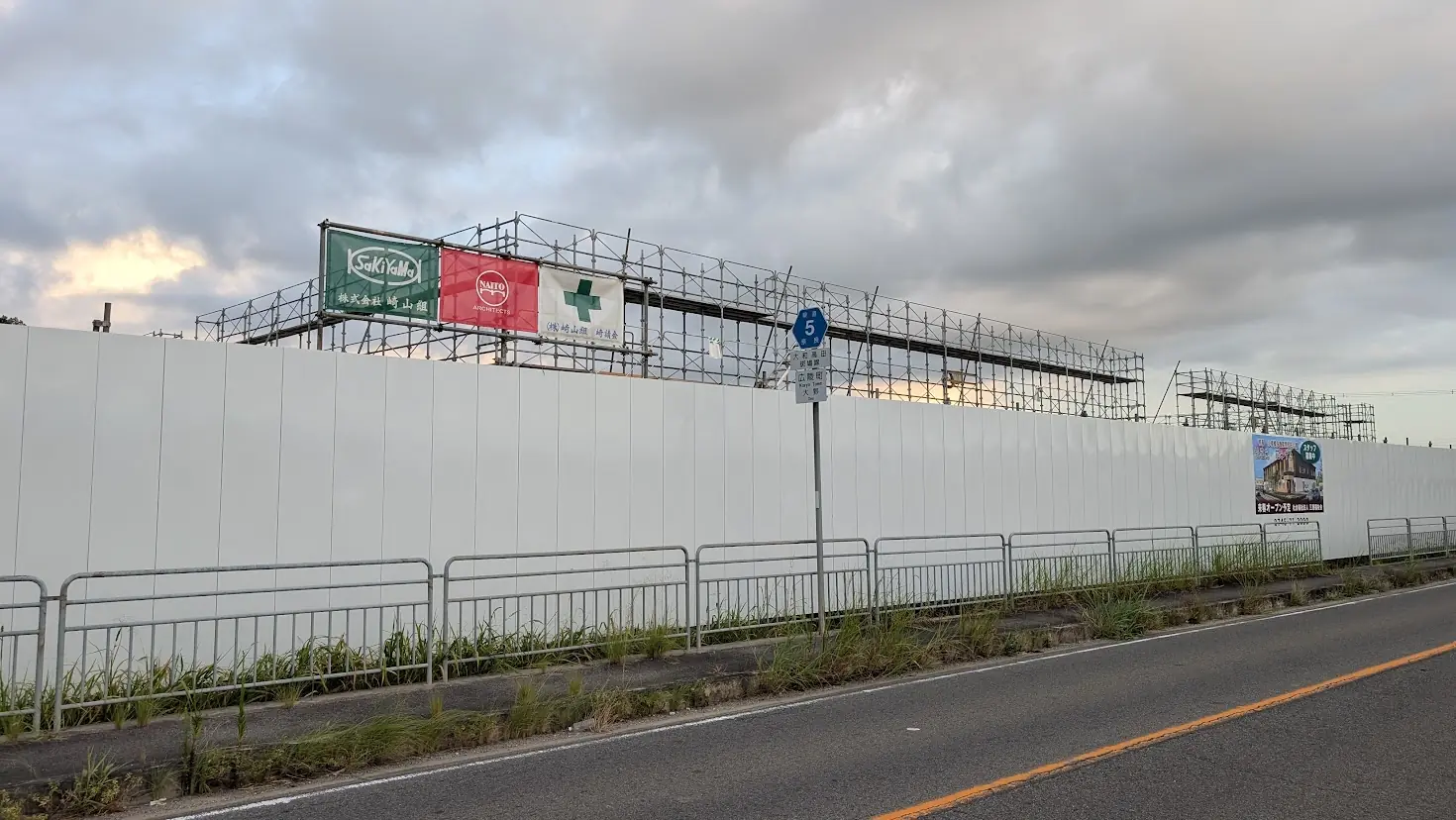 夕暮れの空の下、アスファルト道路の脇に続く白い仮囲いと、「特養 桂花苑」「花みずき」の完成予想図看板が掲げられた建設現場の遠景。