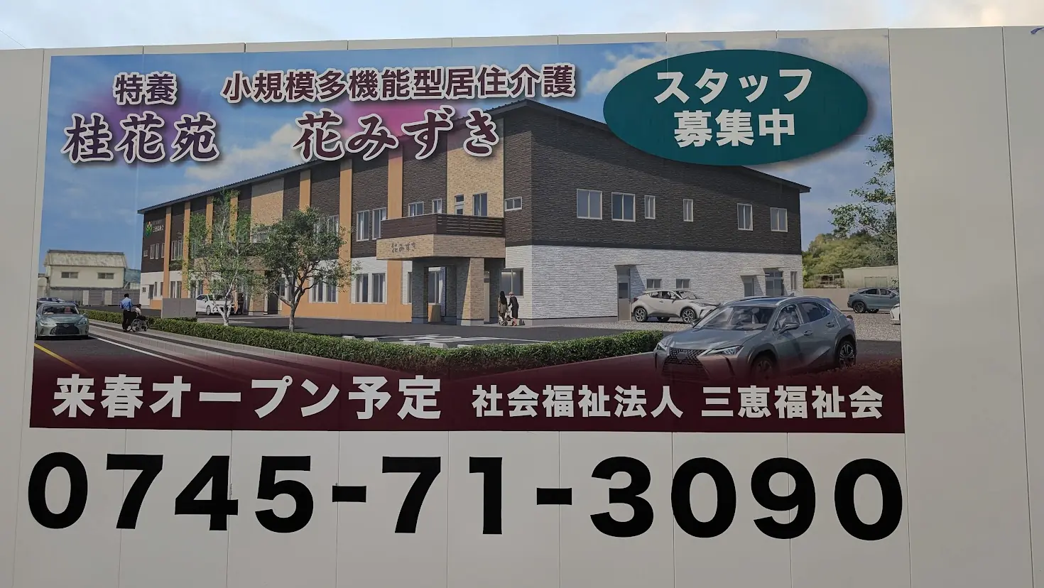 「特養 桂花苑」「小規模多機能型居宅介護 花みずき」の完成予想図と、来春オープン予定の旨・連絡先の電話番号が大きく記載された建設現場看板のアップ。