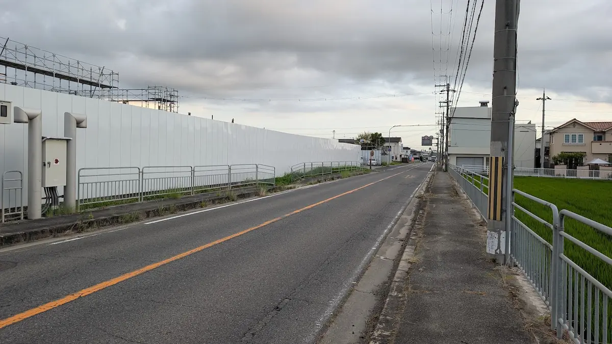 夕暮れの空の下、法隆寺方面へ向かう県道沿いの道路の様子。左側には白い仮囲いに覆われた建設現場のフェンスが続き、右側には田園風景が広がる遠景。