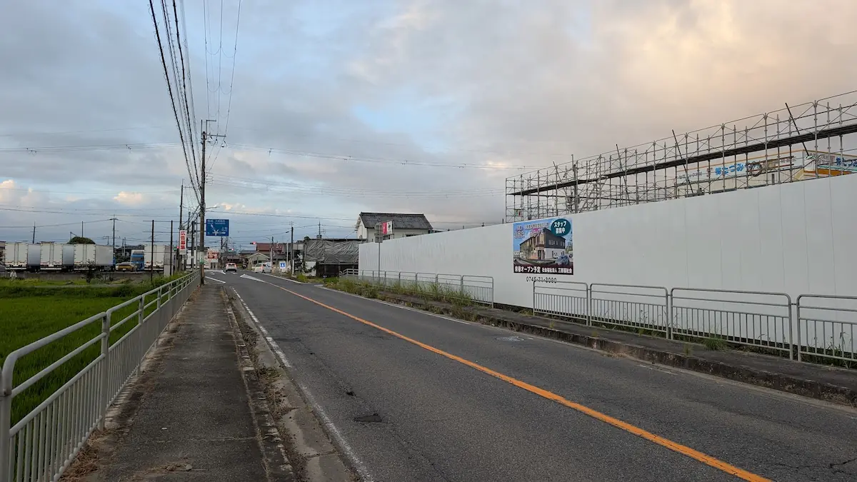 夕暮れの空の下、高田方面へ向かう道路の様子。右側には白い仮囲いに覆われた特養の建設現場があり、奥にローソンの看板が小さく見える遠景。