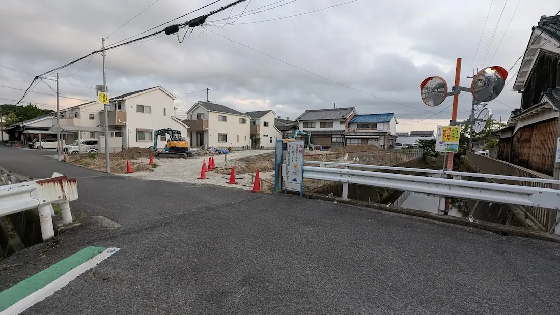 曇天の下、広陵町の住宅街にある道路や橋の脇で進められている工事現場の全体像。重機やカラーコーン、周囲の建物が並ぶ様子。