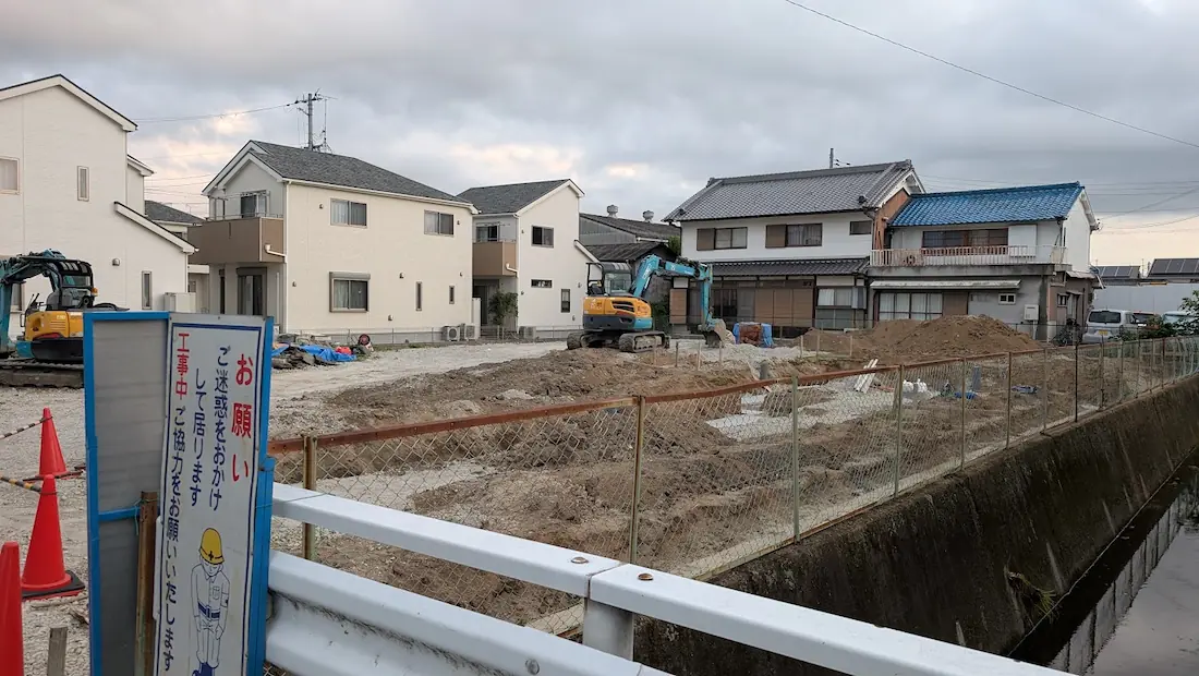 住宅街の川沿いから撮影した工事現場の様子。手前にはガードレールと工事の看板、奥には重機や土砂、民家が並ぶ景色。
