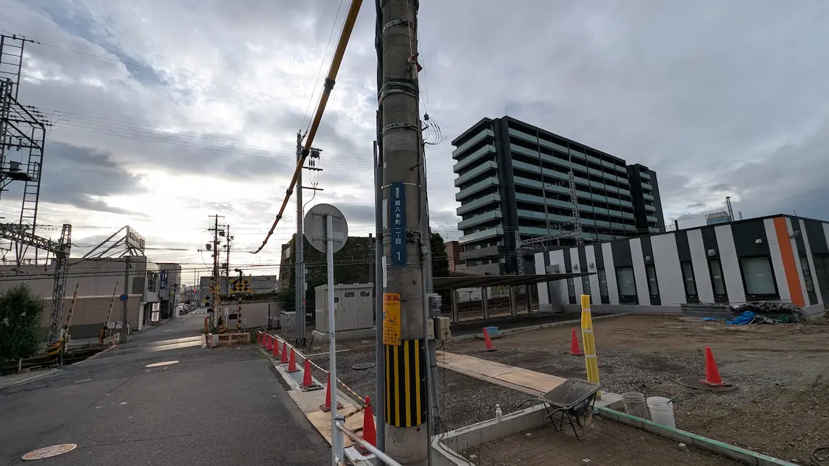 電柱に「南八木町一丁目」の表示札が掲げられ、踏切の先にある道路脇で工事が進められている周辺の様子。