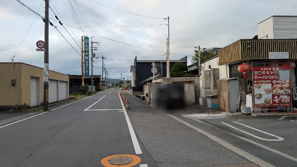 曇天の下、出合交差点を明日香方面へ曲がった先から前方の車道や沿道の建物、駐車車両を見渡した様子。