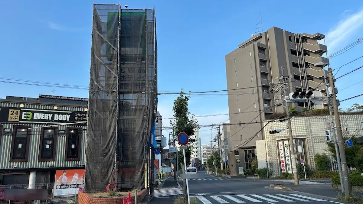 青空の下、閉店した橿原ダイソー付近から新賀北交差点を経由して八木方面へ続く道路の景観。工事中の看板塔や周辺のビルが写っている。