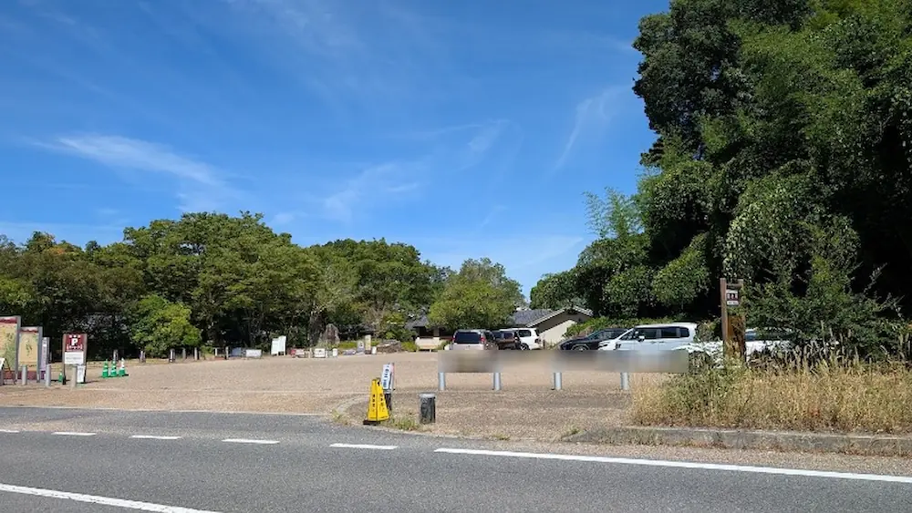 青空の下、緑豊かな木々に囲まれた高松塚古墳の駐車場。誘導スタッフが立ち、数台の車が停められた混雑が落ち着いた駐車場の様子。