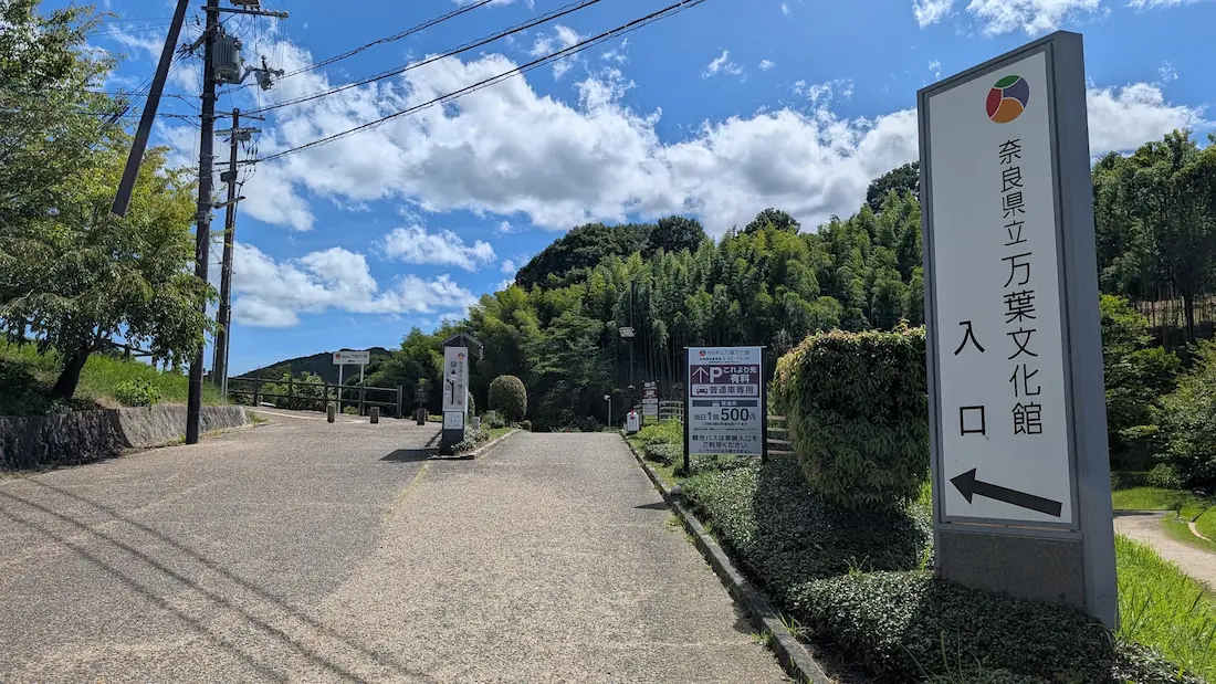 青空の下、奈良県立万葉文化館の駐車場入口に設置された「これより先有料 普通車専用 当日1回500円」と書かれた案内看板と進入路の様子。