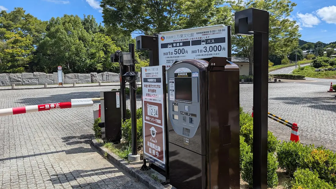青空の下、奈良県立万葉文化館の駐車場に設置された精算機やカメラ、遮断機を備えた自動料金決済システムの全景。
