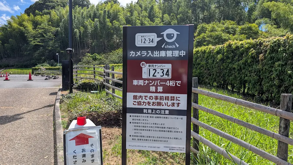 青空の下、奈良県立万葉文化館の駐車場に設置された、「カメラ入出庫管理中」「車両ナンバー4桁で精算」とシステムや館内での事前精算への協力を促す案内看板。